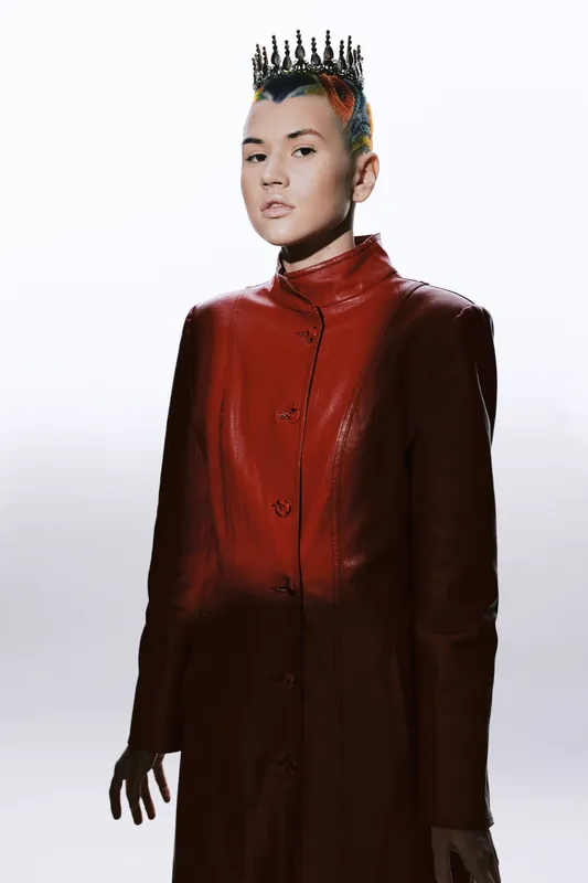 Woman with colorful short hair wearing a red leather coat and a black crown against a bright white background, showcasing modern fashion and fierce attitude.