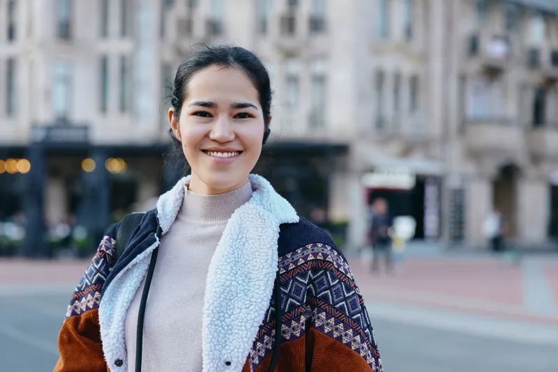 Smiling woman in a warm layered outfit stands on an urban street, wearing a cozy knit sweater, a patterned jacket and scarf, capturing casual city life and everyday style.