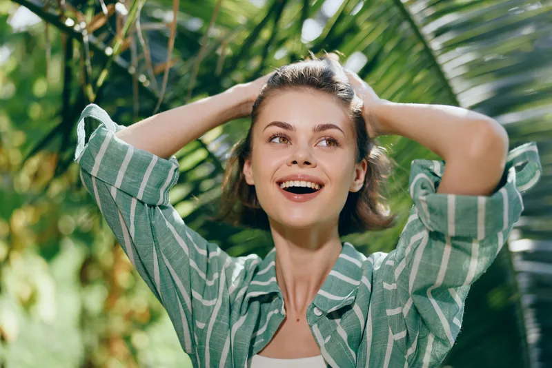 Joyful young woman outdoors with hands in her hair, wearing a green striped shirt, sunlit garden setting.