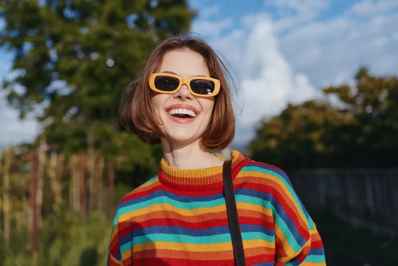 Woman smiling in a colorful striped sweater and yellow sunglasses, short hair portrait outdoors on a sunny day. Casual outfit, joyful expression and playful pose in green park setting for lifestyle.