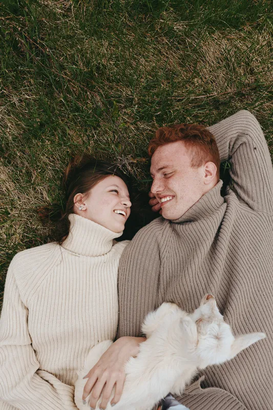 A young couple lies on grass wearing cozy sweaters while holding a small white baby goat, sharing a joyful moment in a natural lifestyle setting outdoors.