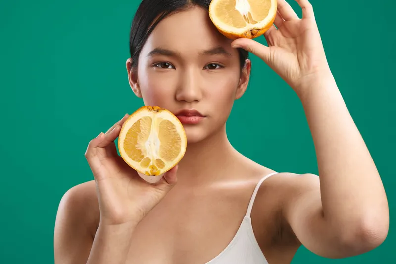 asian woman holds two orange halves against a vivid green backdrop, conveying fresh skincare vibes and playful vitality with a calm, confident gaze.