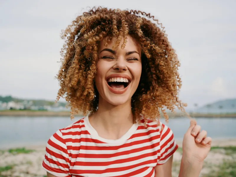 Portrait of a smiling woman with curly hair in a red and white striped shirt, captured outdoors by a lakeside, exuding vibrant energy and natural charm.