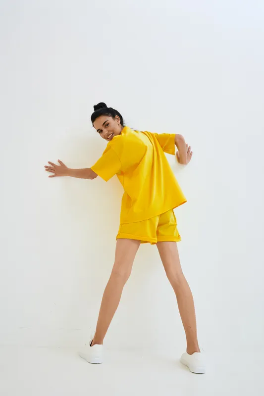 woman in yellow casual outfit standing against white wall, looking back with playful smile, sporty clothing, minimalistic bright studio background, youthful energy