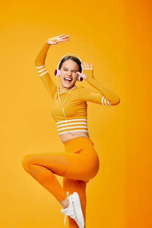 Woman in bright orange fitness outfit dancing mid-air with headphones against a bold yellow backdrop, showcasing energetic movement and athletic style for cardio and studio workouts.