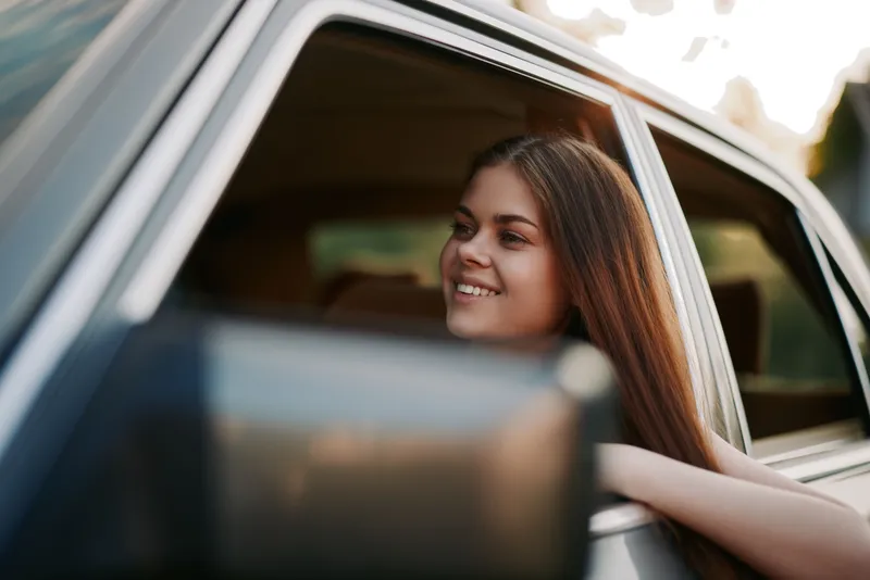 Happy young woman enjoying a road trip, smiling out of a car window, with natural light and a vibrant summer background. Adventure, freedom, joy, travel.