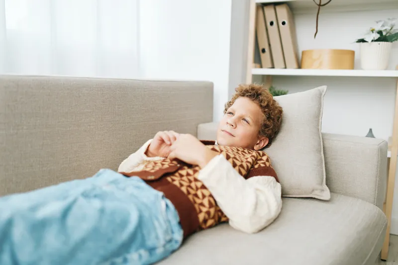 Young boy relaxing on a couch in a cozy living room, wearing a knitted sweater, lounging under a soft blanket with plush pillows and warm light.