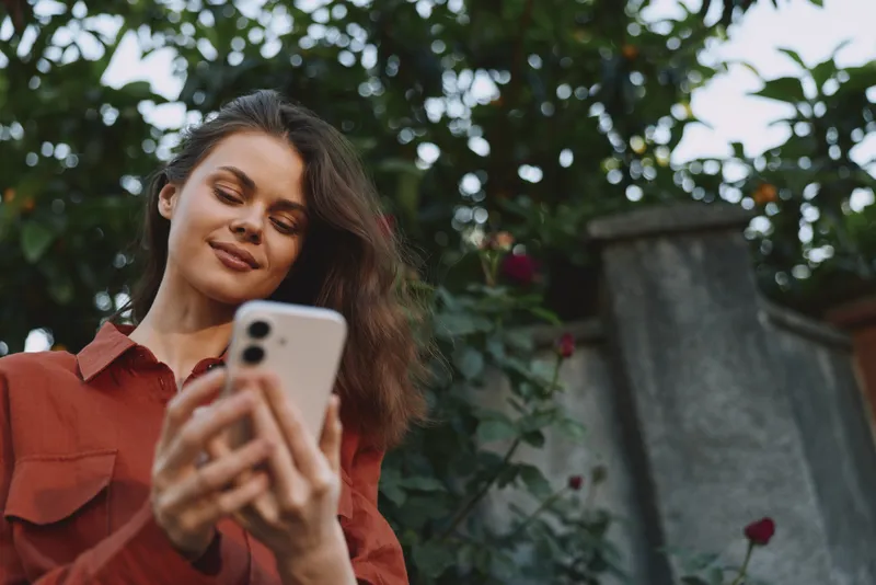 Woman lifestyle outdoor selfie smartphone casual portrait greenery sunlight relaxed, smiling, candid moment with natural colors, red shirt, outdoor background, fresh and approachable vibe