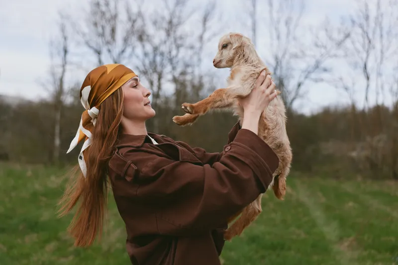 A joyful woman outdoors lifts a small goat kid in a sunlit field, showing playful bonding and rural lifestyle amid calm nature, green grass, and wide sky.