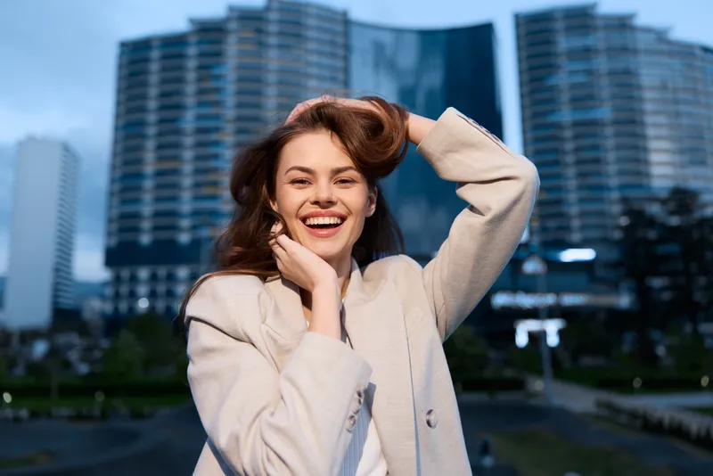 Confident businesswoman smiling outdoors in a contemporary urban setting, wearing a stylish light coat against a modern skyline backdrop.