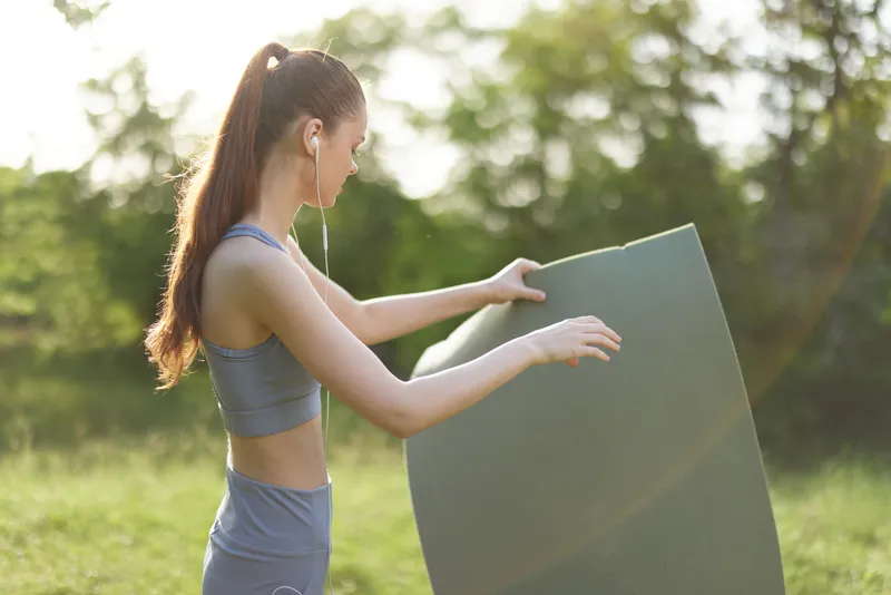 Young athletic woman preparing her yoga mat outdoors, showcasing fitness and wellness in a natural green environment with a calm expression
