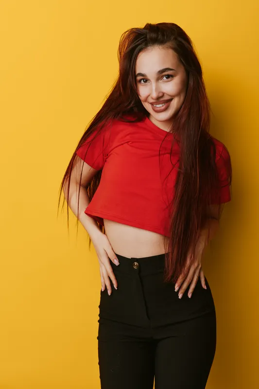 Young woman smiling with long hair wearing red cropped t shirt and black pants standing against yellow background looking confident and happy in studio portrait.