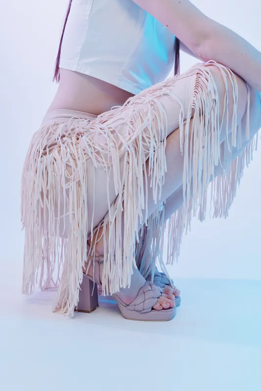 Fashionable woman wearing fringed pants and high heel sandals poses in a studio with soft blue lighting. Concept of beauty, fashion, and modern style.