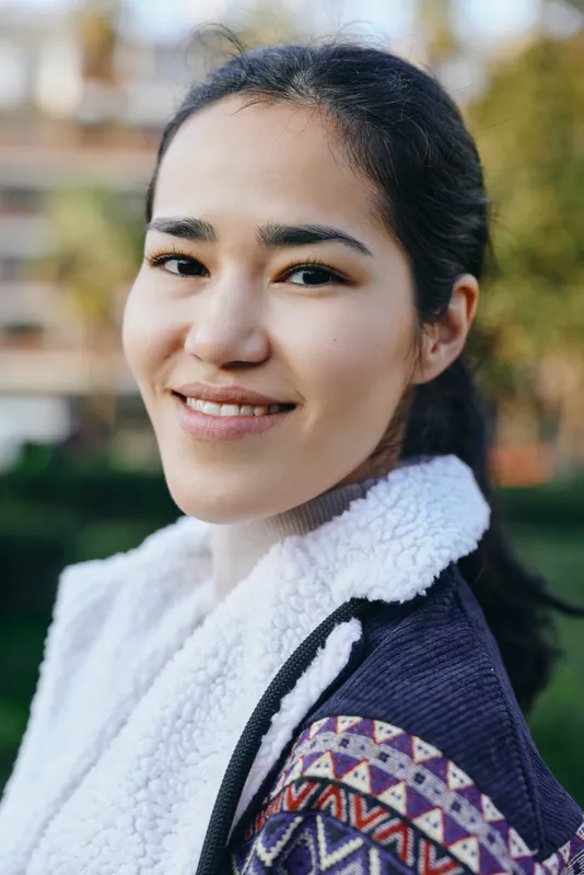 Woman smiling outdoors wearing a cozy scarf and patterned jacket, natural light, soft blurred background, casual fashion shot for lifestyle, outdoor apparel, comfort and warmth in everyday styling