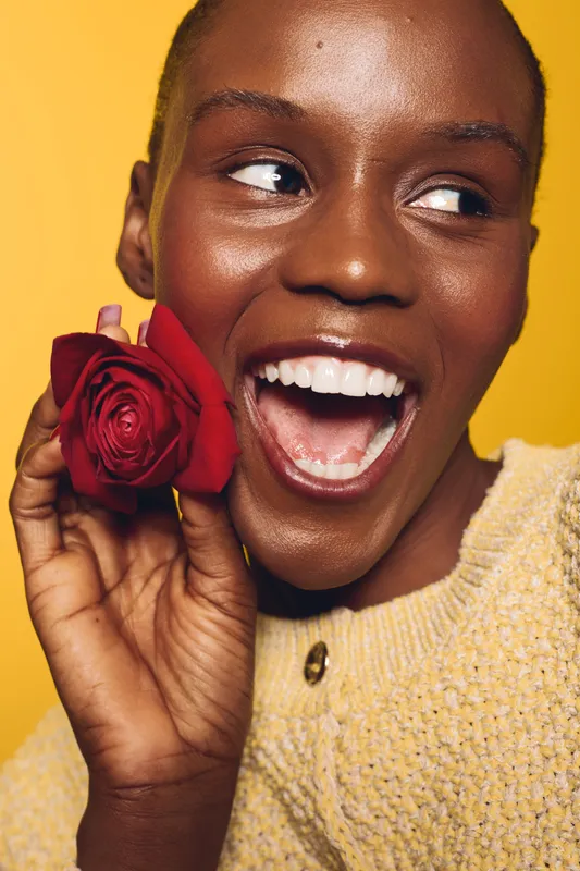 Happy African woman with dark skin holds red rose near face, smiling widely. Yellow background and cozy sweater create warm, joyful mood. Concept of beauty, happiness, and natural elegance.