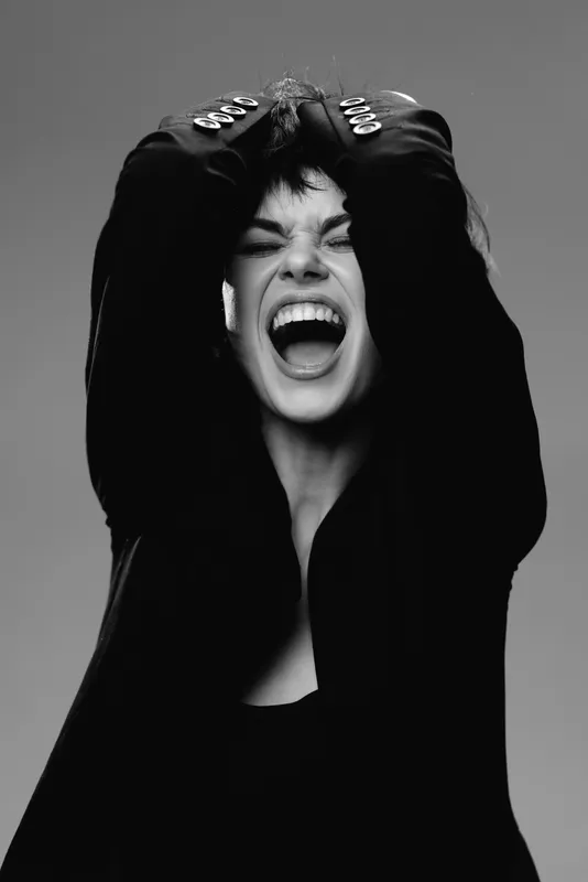Excited woman shouting with joy in black and white, wearing a dark jacket with silver buttons, expressing strong emotion.