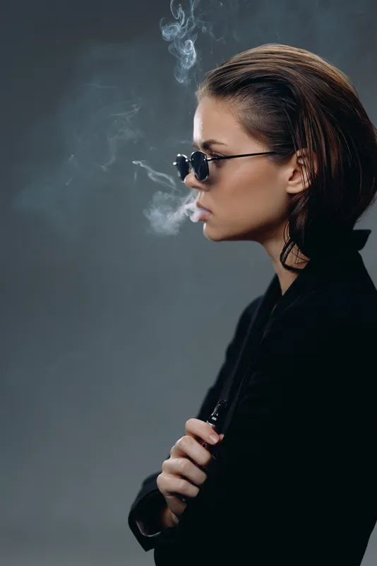 Stylish woman smoking cigarette wearing sunglasses and black jacket with smoky gray background