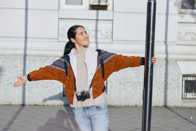 Woman with open arms on a city street, outdoors, wearing a knit sweater and camera at neck, smiling and enjoying freedom, casual urban moment, cheerful vibe