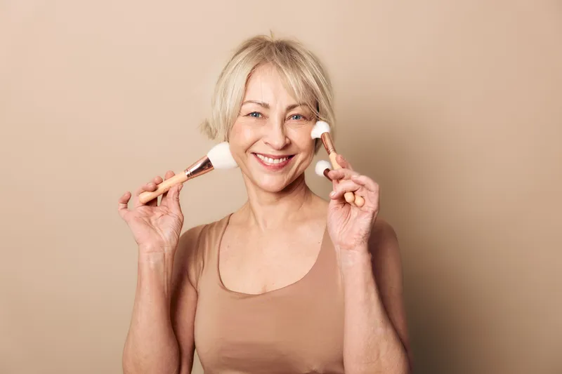 Smiling mature woman with short blonde hair holds two makeup brushes near face on beige background. Concept of beauty, skincare, cosmetics, self-care, and natural aging with positive mood.