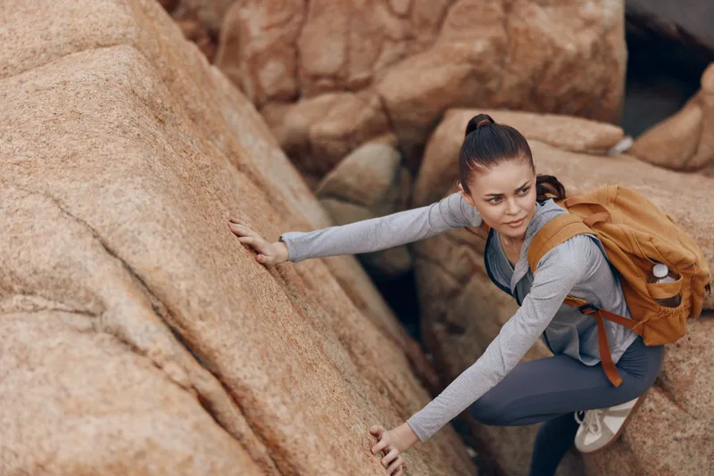 Active young woman climbing rocks, wearing a grey athletic outfit and a brown backpack, showcasing determination and adventure in a natural outdoor setting