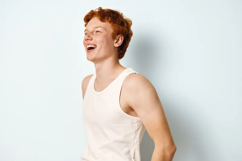 Happy young redhead man laughing and standing in white tank top against plain light background with casual posture and bright mood.