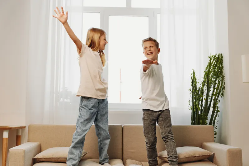 Two happy children standing on a sofa in a bright living room, playing and laughing. The playful boy and girl enjoy a joyful moment with casual clothes and natural light.