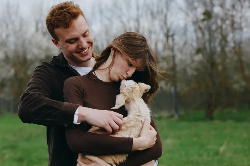 Joyful couple embraces a small goat kid outdoors in a grassy meadow, capturing warmth, bonding, and gentle affection. Natural light enhances a candid moment of countryside lifestyle and tender