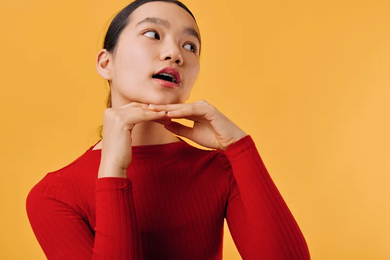 Young woman posing with hands under chin wearing red sweater against yellow background. Female model with natural makeup and dark hair pulled back looking sideways with open mouth.