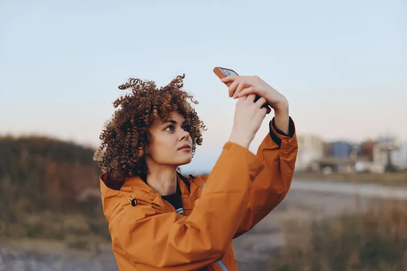 Woman with curly hair in an orange jacket outdoors in autumn, adjusting the hood with her hand, close portrait in a field, nature and sky, lifestyle and leisure
