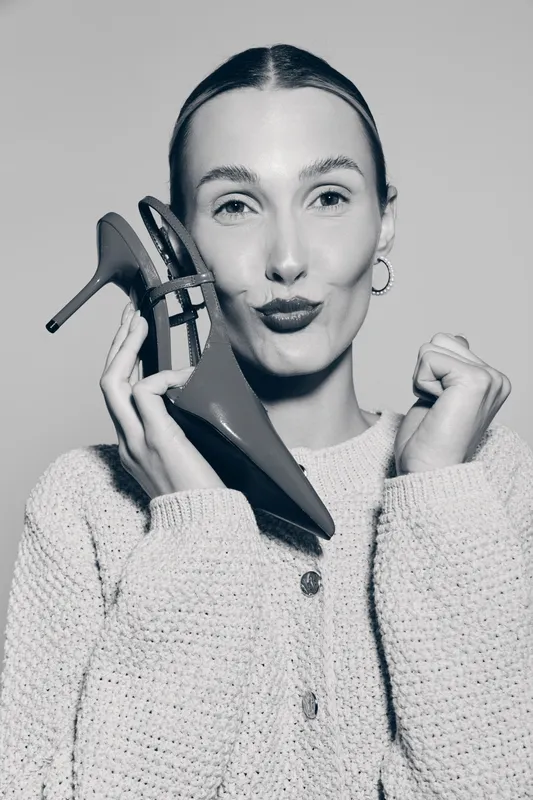 Young white woman with fair skin and slicked hair holds high heel shoe near face and makes a playful expression in studio. Concept of fashion, style, and modern female lifestyle.