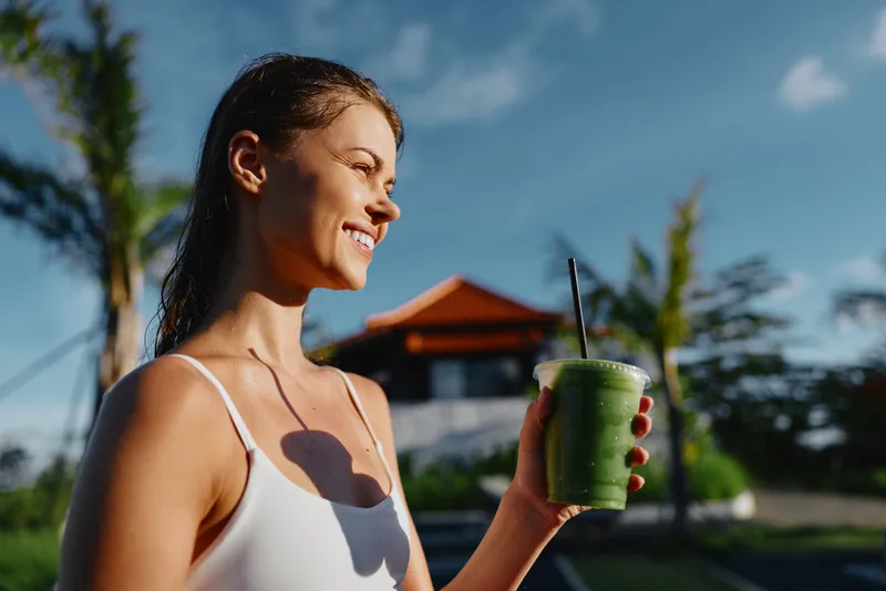 Joyful woman in a white top enjoys a fresh smoothie outdoors, embodying a healthy lifestyle and sunny summer mood with blue sky and plants nearby