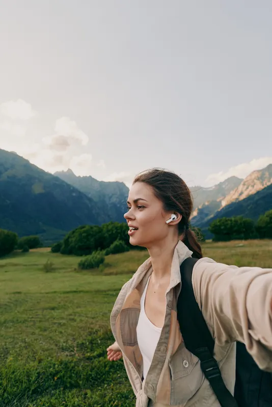 A woman in a light jacket explores a green valley with mountains in the background, conveying an active outdoor lifestyle moment and scenic tranquility.