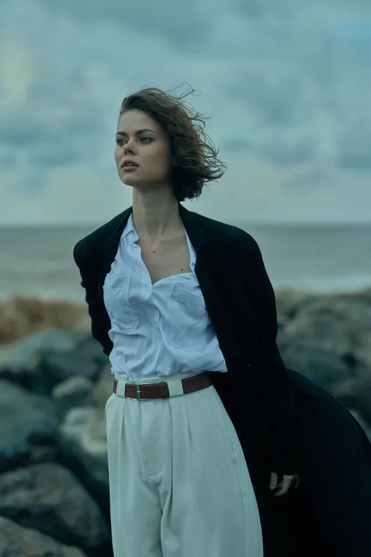 Woman on a windy beach wearing a white blouse and long coat among rugged rocks by the sea, fashion forward mood, outdoor scene.