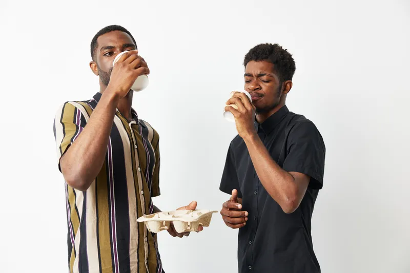 Happy black men enjoying refreshing drinks together, dressed in stylish outfits, sharing a moment of joy in a bright, minimalistic setting with soft lighting.