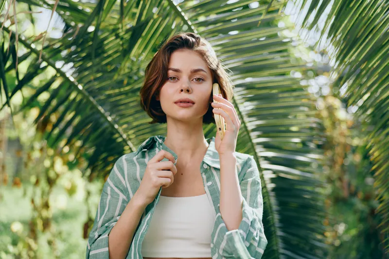 A woman stands outdoors among tropical greenery, wearing a striped green shirt over a white top, gazing softly at the camera with a calm, confident expression.