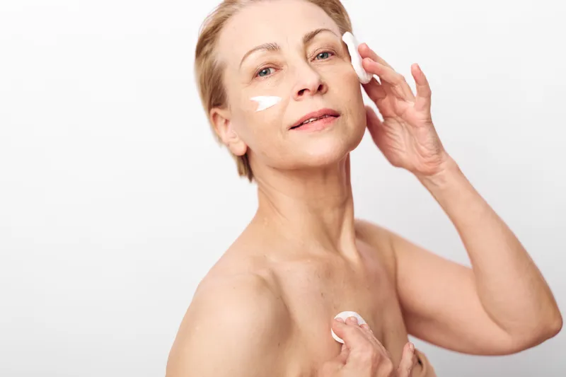 Mature woman with fair skin applies moisturizing cream on face and neck in bright studio. Concept of skincare, anti aging, beauty routine, and natural self care for healthy skin.