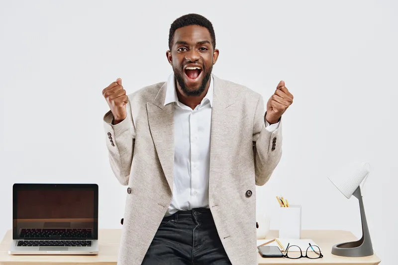 Confident young man with dark skin expressing enthusiasm, standing in an office environment with a laptop, white desk lamp, and glasses, showcasing success and positive emotions. People lifestyle conc