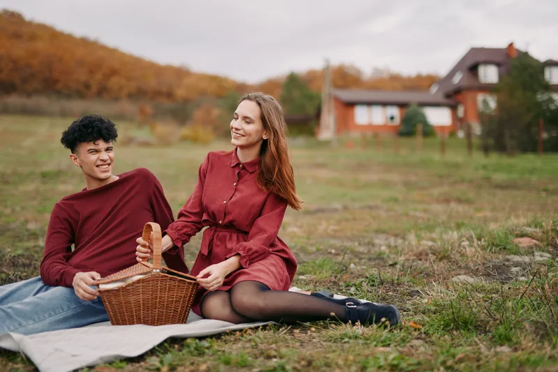 Young couple enjoying a picnic in the countryside, candid expressions and natural poses conveying authenticity and accuracy of a relaxed autumn outing with a wicker basket on a blanket.