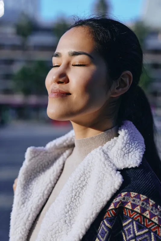 Sunlit moment with closed eyes, a calm woman enjoy warmth outdoors, wearing cozy coat and patterned jacket, serene mood, gentle breeze, soft shadows and morning glow