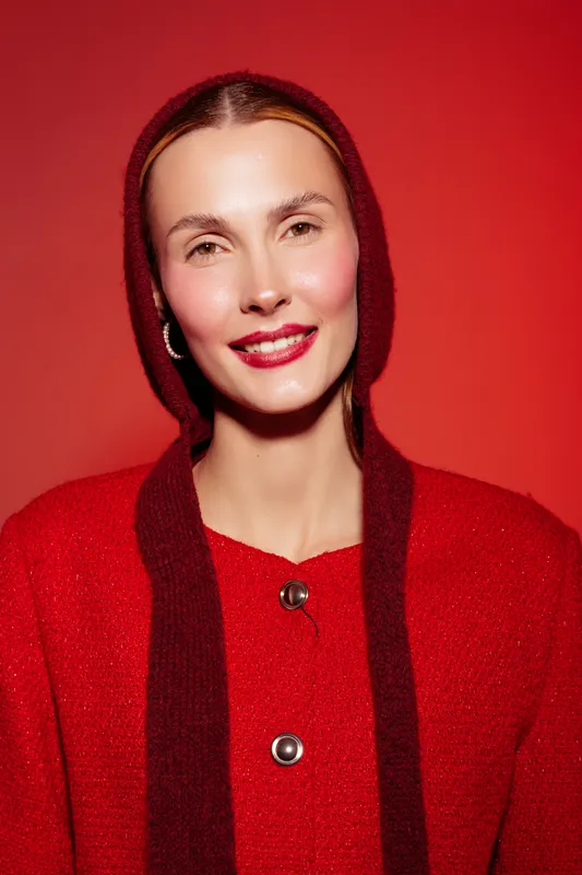 Smiling woman in red knitwear with hood poses against red background. Close-up portrait with natural makeup and hoop earrings. Concept of fashion, style, and winter clothing.
