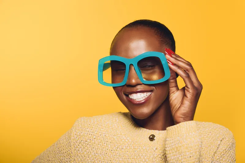 Smiling dark skinned African American woman wearing oversized blue sunglasses and yellow sweater poses against bright yellow background. Concept for fashion, joy, and summer style.
