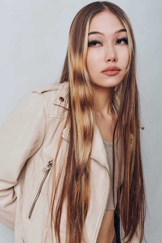 Young woman with long straight hair wearing light beige leather jacket posing with neutral expression on light gray background for fashion beauty portrait.