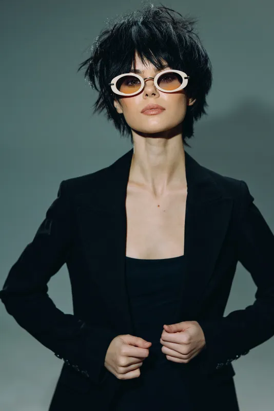 Stylish woman with short black hair and sunglasses wearing a black blazer posing confidently against gray background.