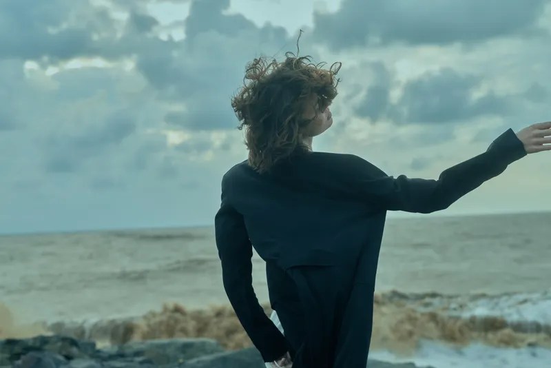 Outstretched arms silhouette on a windy seashore, a dark coat flutters as waves crash, conveying freedom, mystery and resilience in a moody coastal scene.