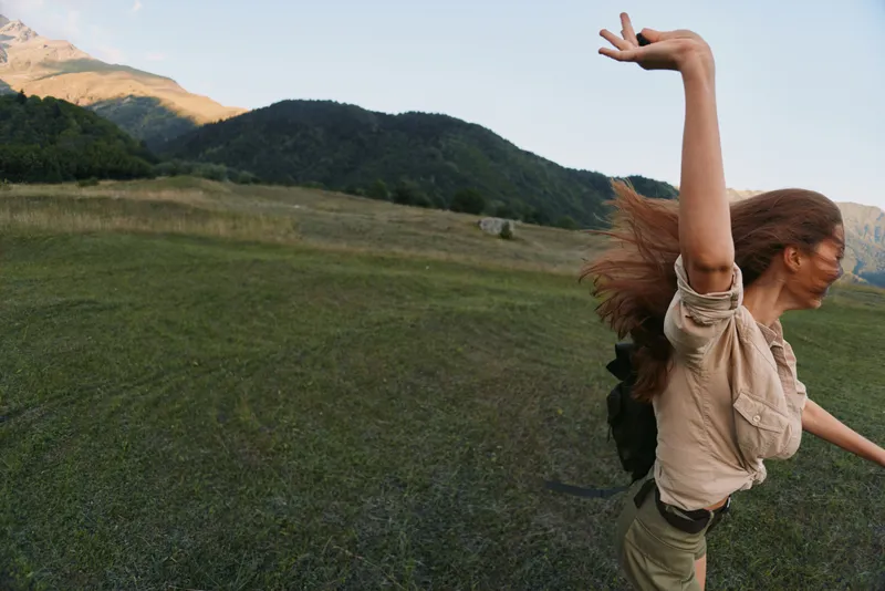 Woman in casual outfit dances in an outdoors field, wind in her hair, embracing a carefree lifestyle under an open sky with distant mountains.
