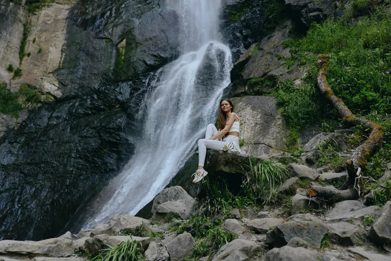 Young woman sitting near a large waterfall surrounded by rocks and lush green vegetation in serene outdoor nature setting.