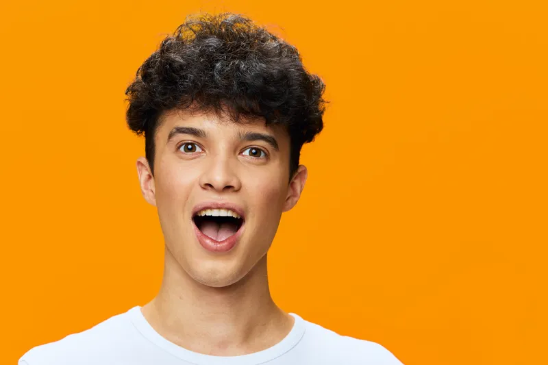 surprised young man with curly hair wearing white shirt stands against bright orange background showing open mouth expression of shock and amazement