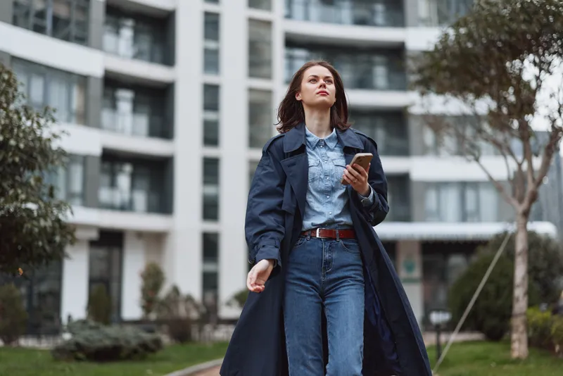 Woman outdoor fashion urban denim coat smartphone confidence posing street, looking up with a calm gaze in a sunlit moment, modern architecture in the background