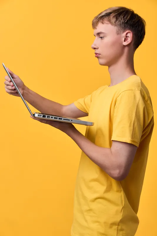 Young man in a yellow shirt using a laptop against a bold orange background, illustrating remote work, digital devices, and modern productivity in a studio scene