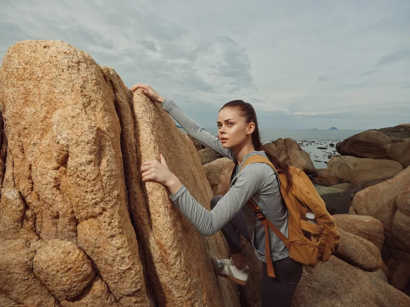 Young female hiker climbing rocks with a backpack near the sea, showcasing adventure and determination in an outdoor summer setting